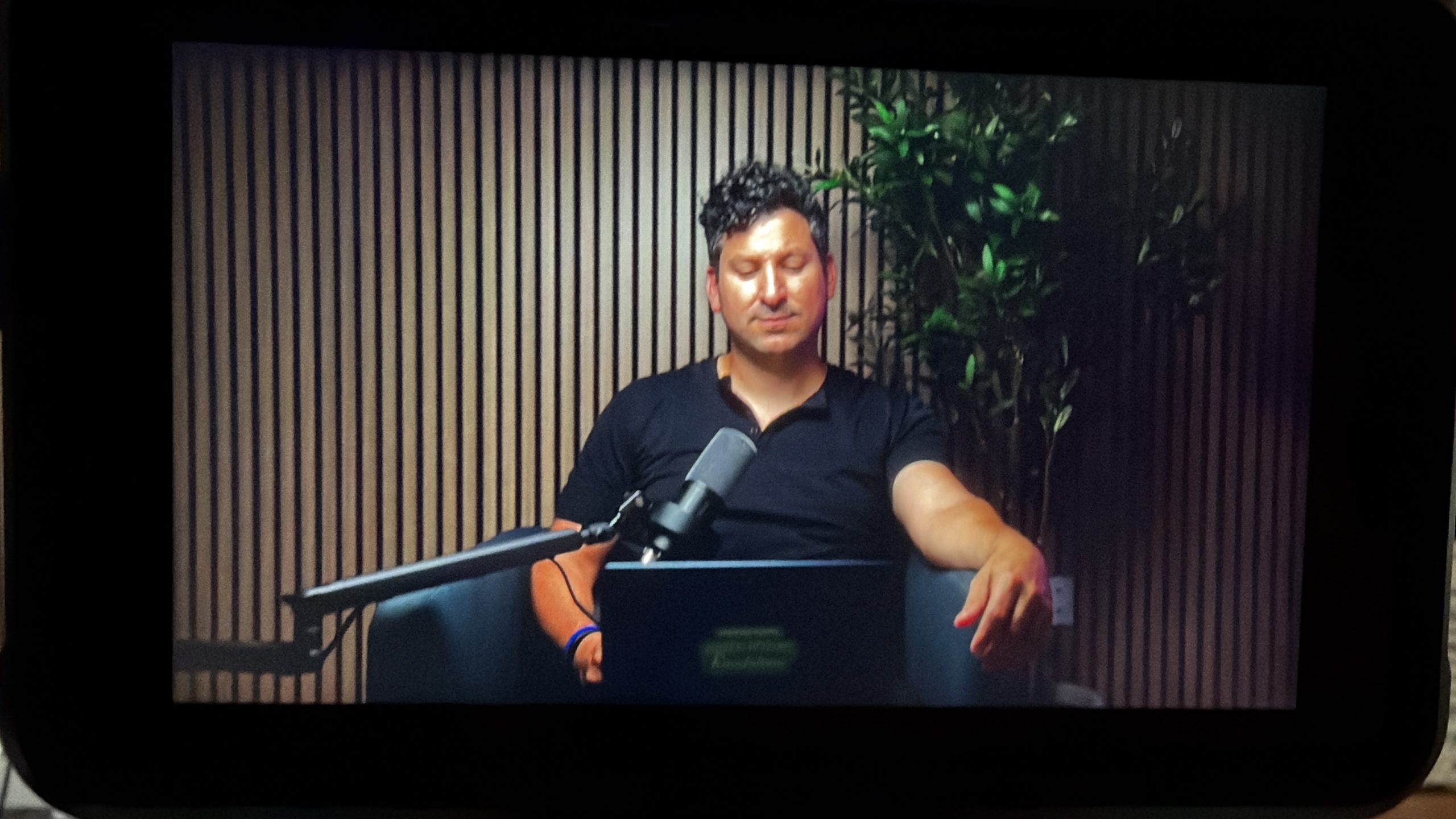 Monitor showing a man recording a podcast episode at a microphone in front of the wood slat wall