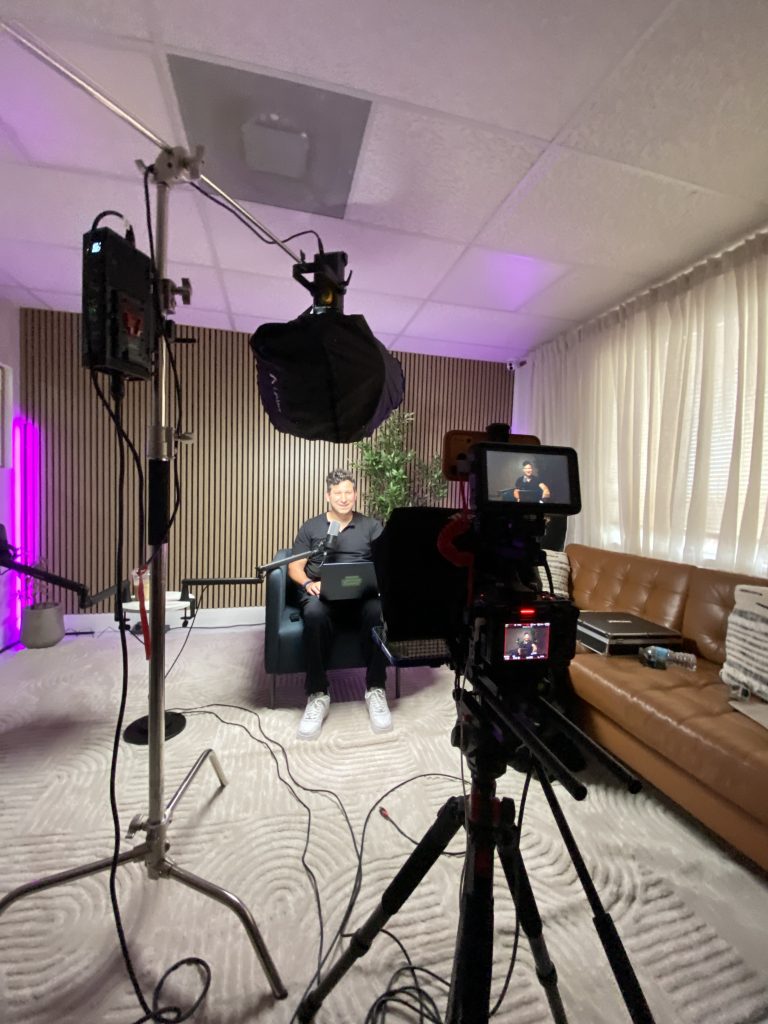 Podcast host smiling on set with a laptop while the camera monitor shows the live framing