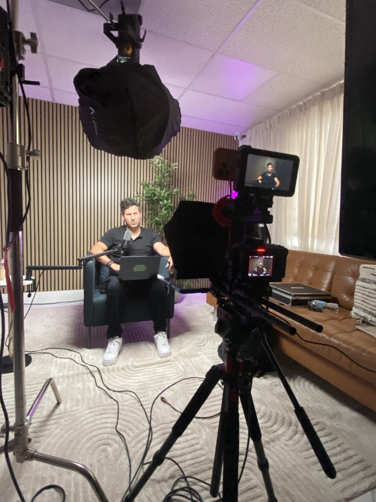 Camera with two monitors recording a podcast host working from a laptop on the studio set