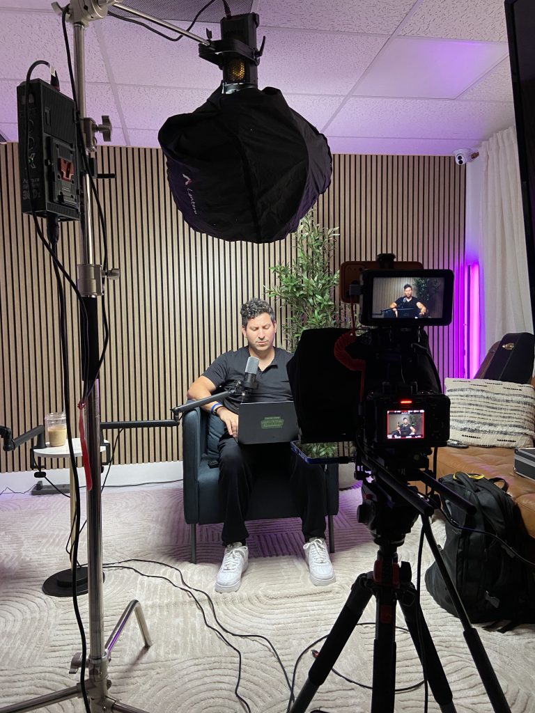 Podcast host reading from a teleprompter laptop while a camera monitor shows the live shot