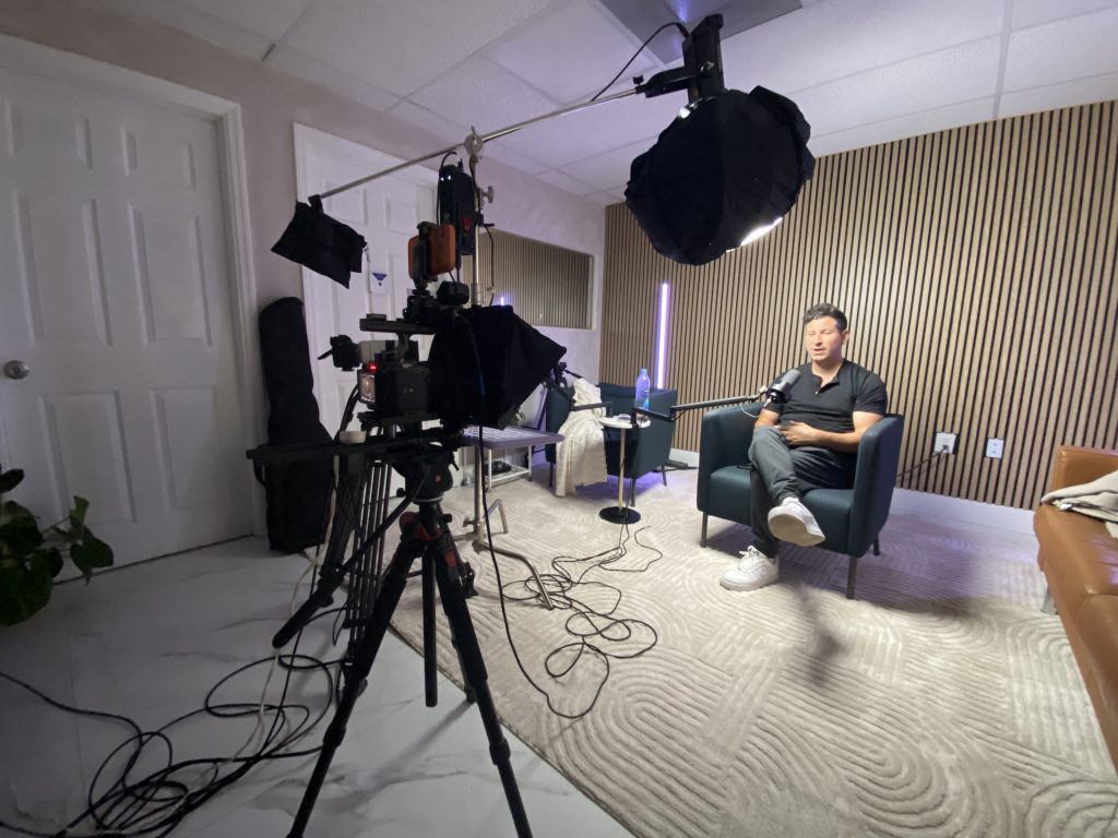 Podcast studio set with a camera on a tripod at left and the host seated under studio lighting at right