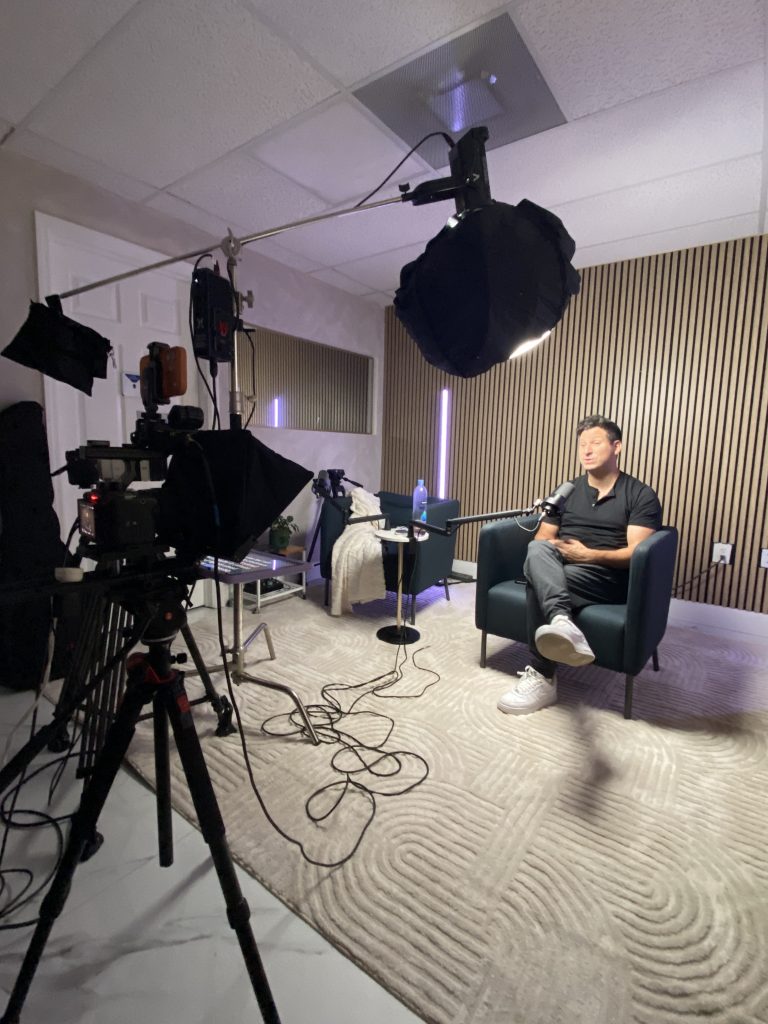 Cinema camera on a tripod framing a podcast host seated in an armchair under a boom-mounted softbox