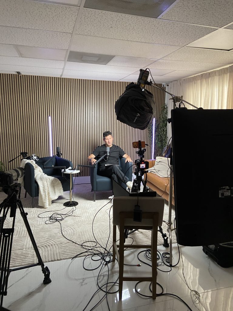 Podcast set seen from beside the monitor, with the host at the microphone and a camera rig in the foreground