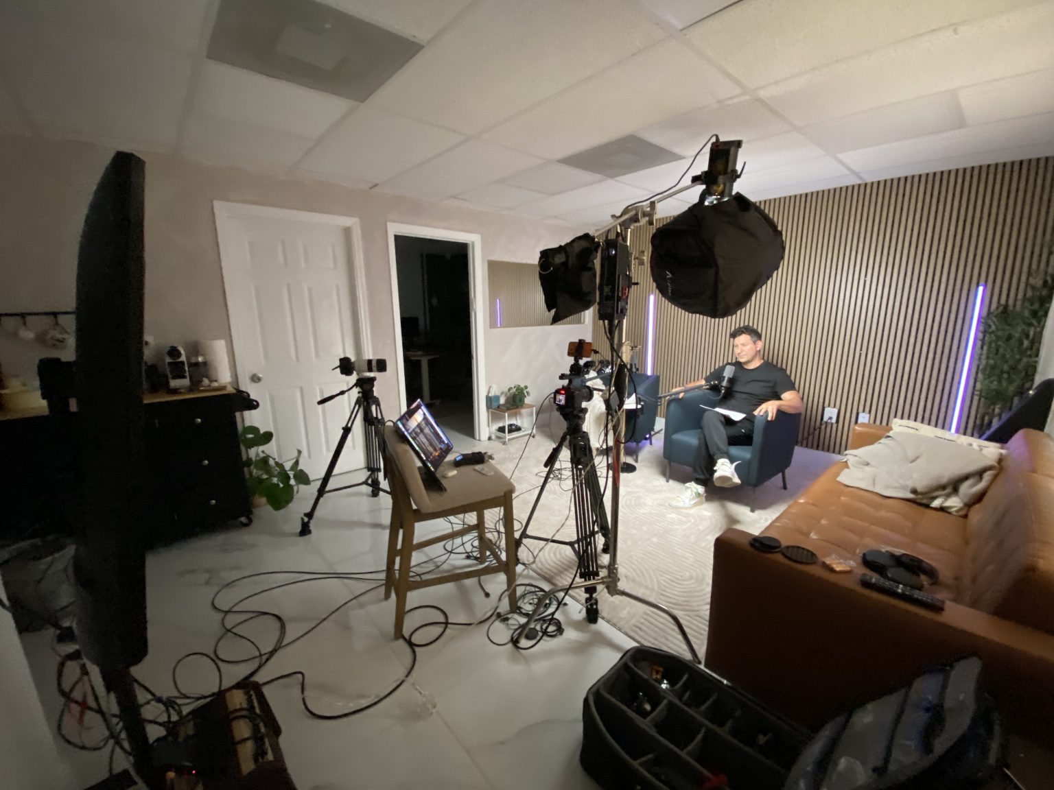 Wide view of the full podcast rig: two cameras on tripods, an overhead softbox, an acoustic flag and a laptop on a stool