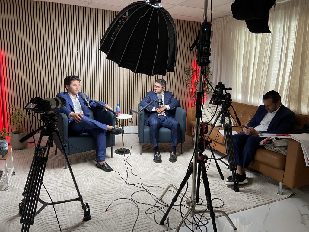 Two podcast guests at microphones facing each other in the studio armchairs, with cameras and a softbox rigged around them