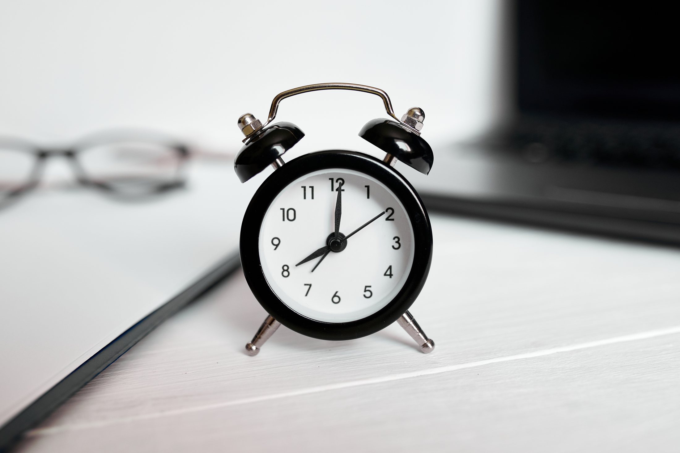 Black alarm clock on a desk beside glasses and a laptop